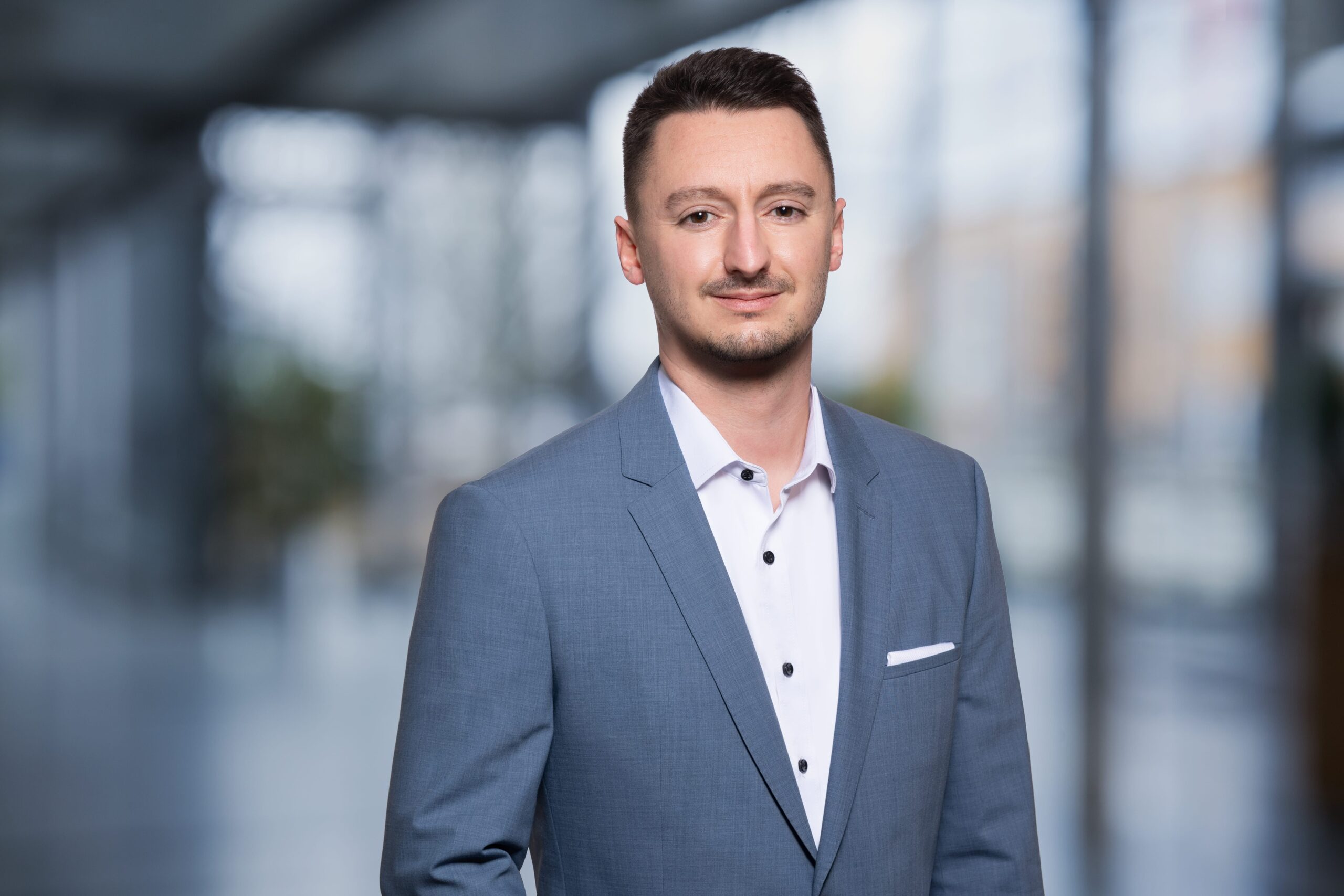 558747987 3750f965 A0a4 4722 9907 92a05d3efbce Mann in grauem Anzug mit weißem Hemd, steht in moderner Umgebung mit unscharfem Hintergrund.