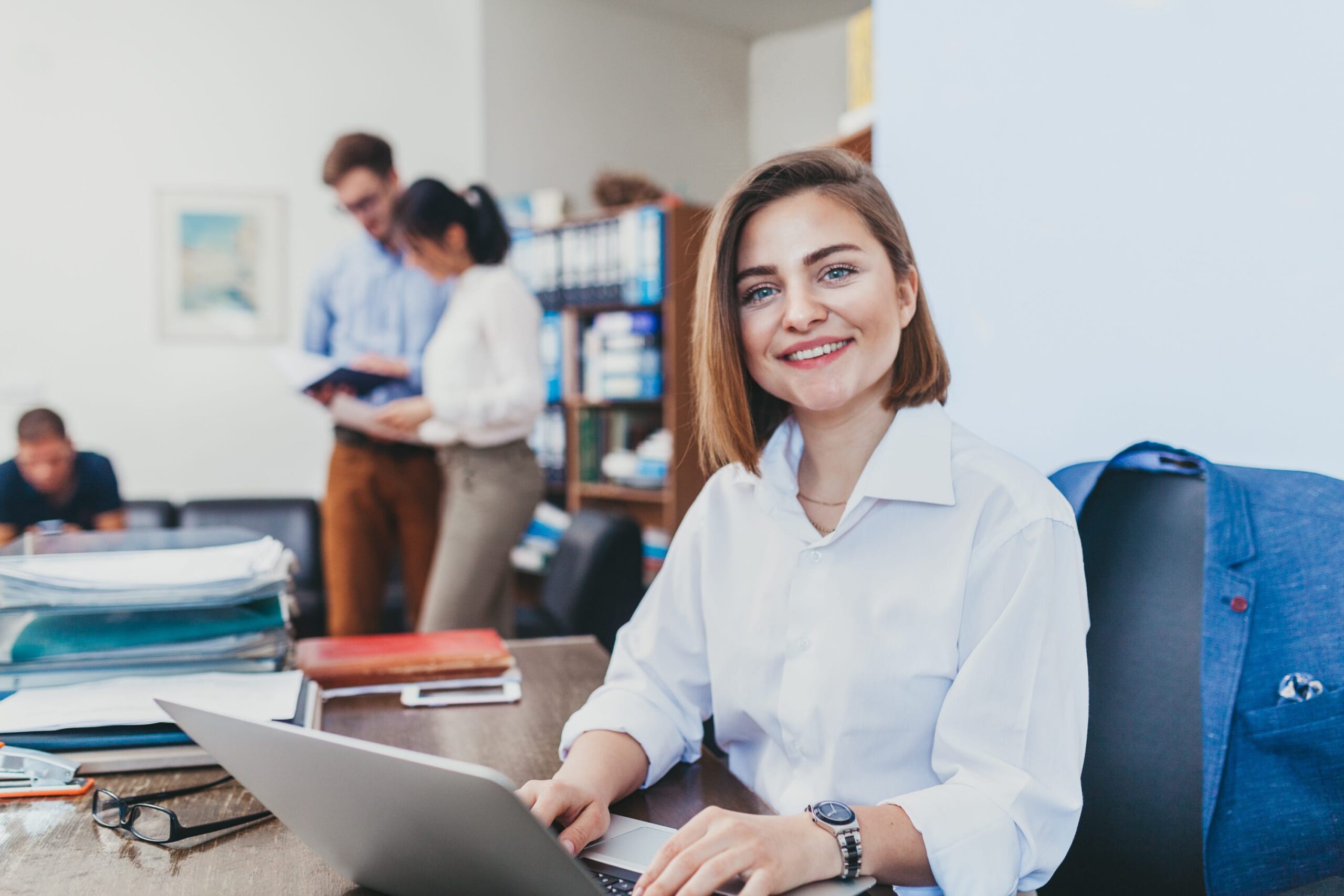 AdobeStock 166883410 Junge Frau mit Laptop in einem Büro, lächelnd, während Kollegen im Hintergrund diskutieren.
